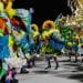 Integrantes de escola de samba desfilam no Carnaval 2026 com fantasias coloridas e bandeiras do Brasil.