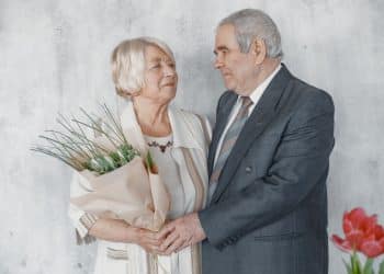 Casal de idosos em trajes formais de casamento. O homem veste um terno cinza e a mulher segura um buquê de flores brancas. Eles estão de frente um para o outro em um fundo claro.