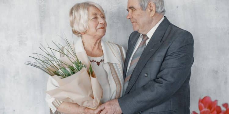Casal de idosos em trajes formais de casamento. O homem veste um terno cinza e a mulher segura um buquê de flores brancas. Eles estão de frente um para o outro em um fundo claro.