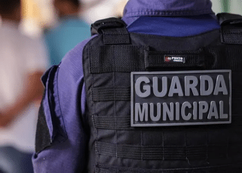 Close no colete tático preto com placa da Guarda Municipal em uniforme azul.