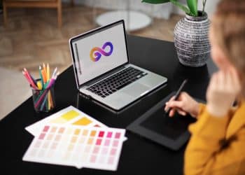 Vista superior de uma designer trabalhando em um laptop que exibe um logotipo colorido em formato de infinito, com uma mesa digitalizadora, amostras de cores e lápis ao redor.