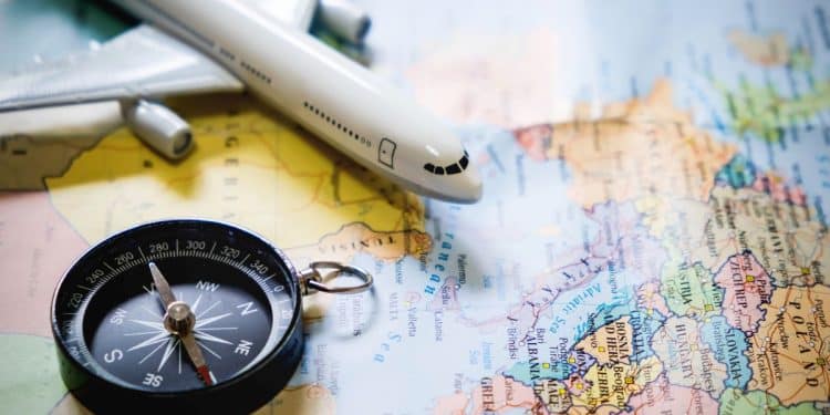 Visão aérea de um mapa-múndi colorido sobre o qual repousam uma bússola preta clássica e a miniatura de um avião comercial branco decolando sobre a Europa e o Mediterrâneo.