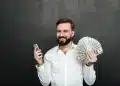 Homem sorrindo enquanto segura um maço de dinheiro e um celular, simbolizando a conquista de um bom salário com oportunidades que não exigem diploma superior.