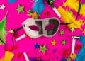 Uma máscara de Carnaval prateada e preta centralizada sobre um fundo rosa vibrante. Ao redor, há estrelas coloridas, penas verdes e amarelas, e apitos de festa espalhados.