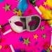 Uma máscara de Carnaval prateada e preta centralizada sobre um fundo rosa vibrante. Ao redor, há estrelas coloridas, penas verdes e amarelas, e apitos de festa espalhados.
