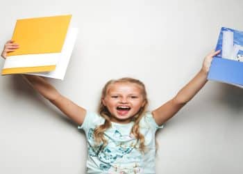 Garota sorridente segurando pastas azul e amarela com objetos escolares.