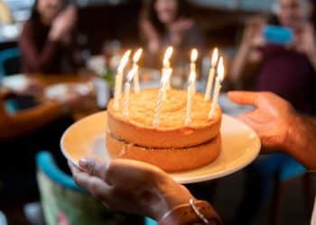 Pessoa segurando um bolo de aniversário com velas acesas, celebrando com amigos em um ambiente descontraído.