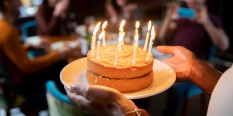 Pessoa segurando um bolo de aniversário com velas acesas, celebrando com amigos em um ambiente descontraído.