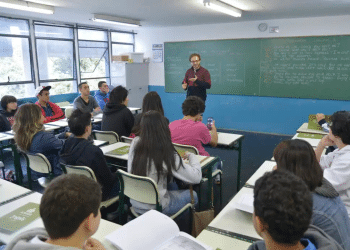 Professor interage com alunos atentos em sala de aula, com quadro negro ao fundo