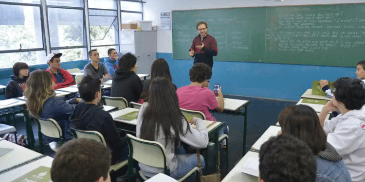 Professor interage com alunos atentos em sala de aula, com quadro negro ao fundo