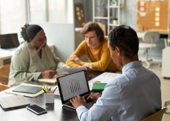 Três profissionais em uma reunião estratégica de negócios; um homem em primeiro plano analisa gráficos de barras em um notebook enquanto conversa com colegas ao fundo.