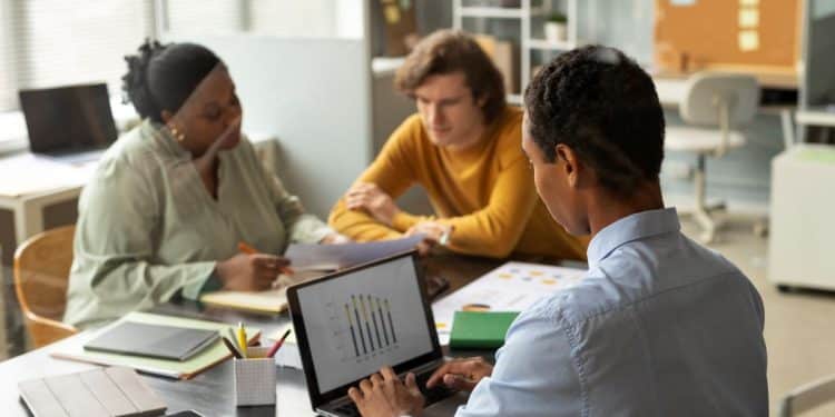 Três profissionais em uma reunião estratégica de negócios; um homem em primeiro plano analisa gráficos de barras em um notebook enquanto conversa com colegas ao fundo.