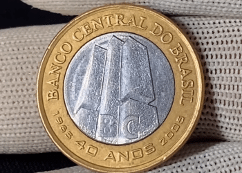 Close-up da moeda de 1 real de 2005, comemorativa dos 40 anos do Banco Central, segurada por luvas brancas. O foco está no detalhe da "data marcada" à direita do edifício central.