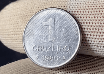 Close de uma moeda prateada de 1 Cruzeiro do ano 1980 sendo segurada por dedos com luvas de proteção brancas.