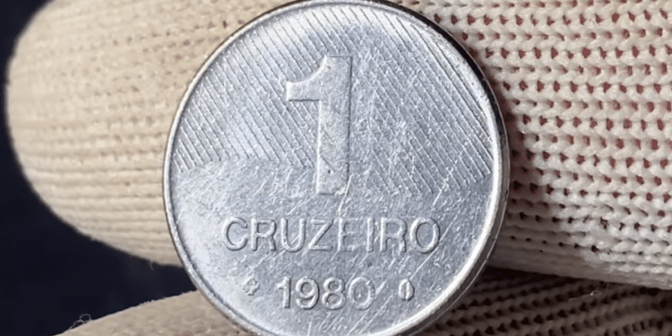 Close de uma moeda prateada de 1 Cruzeiro do ano 1980 sendo segurada por dedos com luvas de proteção brancas.