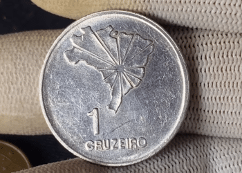 Close-up do reverso de uma moeda prateada de 1 Cruzeiro. No centro, o mapa do Brasil com linhas radiais partindo de Brasília; abaixo, o numeral "1" e a palavra "CRUZEIRO". A moeda é segurada por dedos usando luvas brancas de proteção.