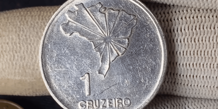 Close-up do reverso de uma moeda prateada de 1 Cruzeiro. No centro, o mapa do Brasil com linhas radiais partindo de Brasília; abaixo, o numeral "1" e a palavra "CRUZEIRO". A moeda é segurada por dedos usando luvas brancas de proteção.