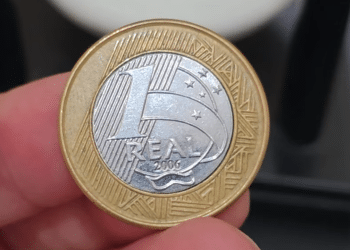 Close-up de dedos segurando uma moeda de 1 real de 2006. O núcleo prateado está visivelmente deslocado para a esquerda, deixando uma abertura no anel dourado.