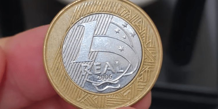 Close-up de dedos segurando uma moeda de 1 real de 2006. O núcleo prateado está visivelmente deslocado para a esquerda, deixando uma abertura no anel dourado.