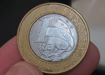 Close-up de uma moeda de 1 real de 2004 sendo segurada por dedos, destacando a data "2004" no centro prateado e as bordas douradas.