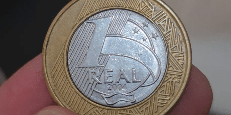 Close-up de uma moeda de 1 real de 2004 sendo segurada por dedos, destacando a data "2004" no centro prateado e as bordas douradas.