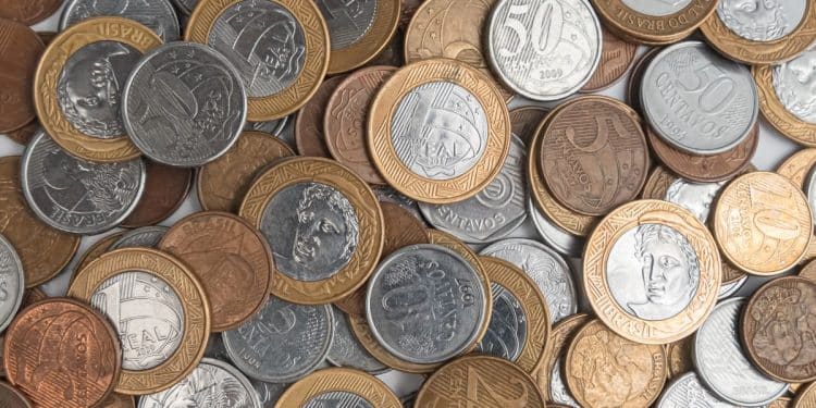 Fotografia em plano fechado de um monte de moedas de Real espalhadas, incluindo moedas de 1 Real, 50 centavos, 10 centavos e 5 centavos. As peças apresentam diferentes anos de cunhagem e estados de conservação, com destaque para o brilho do metal e detalhes do relevo.