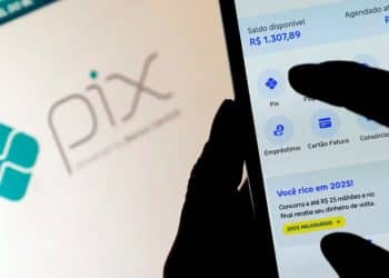 Foto em close de uma pessoa utilizando um smartphone para acessar o aplicativo de um banco, com o logotipo do Pix visível ao fundo em uma tela de computador.