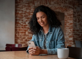 Mulher jovem de cabelos cacheados sorrindo enquanto olha para a tela de um smartphone em um café, com fundo de tijolos aparentes desfocado.