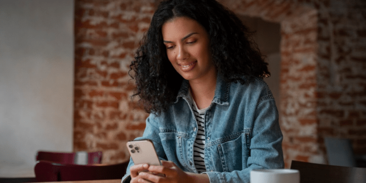 Mulher jovem de cabelos cacheados sorrindo enquanto olha para a tela de um smartphone em um café, com fundo de tijolos aparentes desfocado.