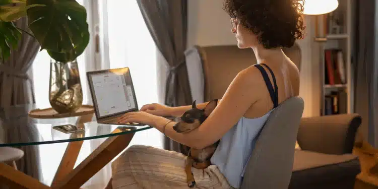 Mulher trabalhando de casa com um cachorro no colo, usando um laptop enquanto organiza suas tarefas em um ambiente aconchegante.