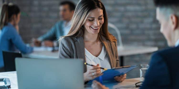 Executiva sorridente analisando documentos em reunião de trabalho com notebook e colegas ao fundo.