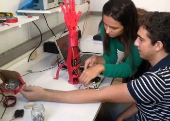 Duas pessoas montando uma prótese de mão robótica vermelha em laboratório de engenharia biomédica