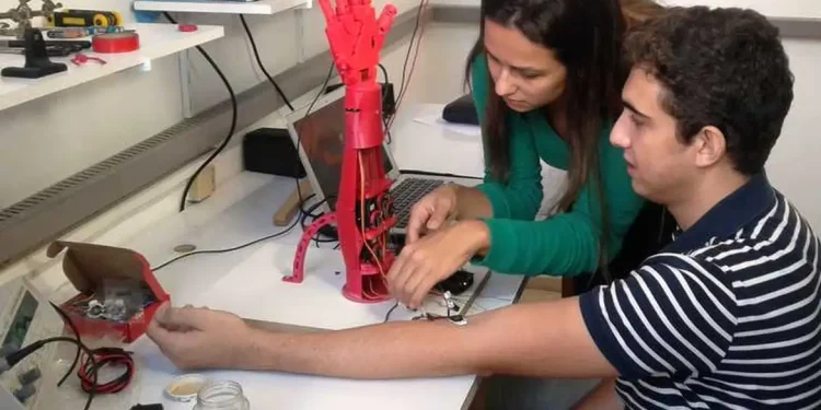 Duas pessoas montando uma prótese de mão robótica vermelha em laboratório de engenharia biomédica
