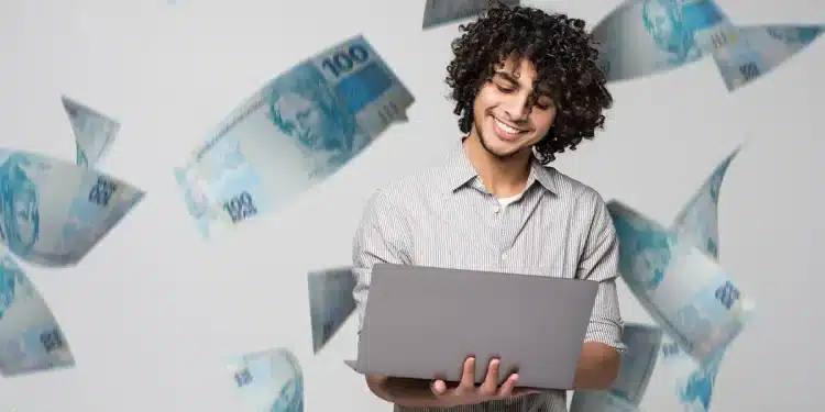 Homem sorrindo enquanto usa laptop, com notas de R$ 100 flutuando ao redor, simbolizando sucesso financeiro.