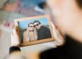 Visão em primeira pessoa de alguém segurando um porta-retrato de madeira com a foto de um casal sorridente à beira de um lago. O foco está na moldura, enquanto o fundo permanece desfocado.