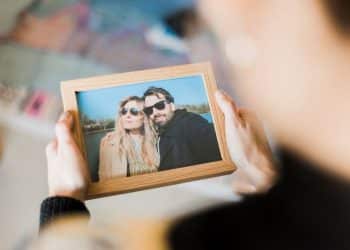 Visão em primeira pessoa de alguém segurando um porta-retrato de madeira com a foto de um casal sorridente à beira de um lago. O foco está na moldura, enquanto o fundo permanece desfocado.
