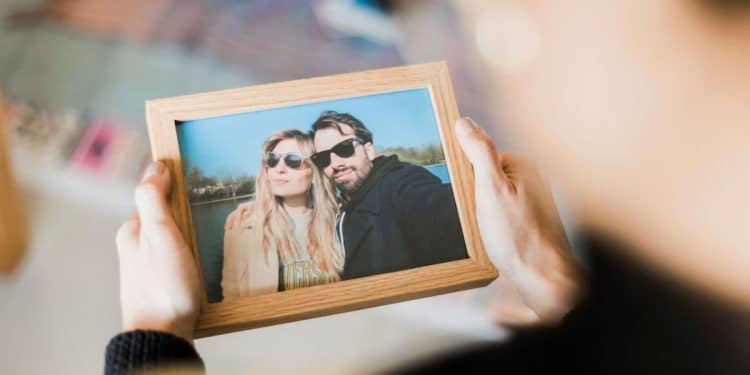 Visão em primeira pessoa de alguém segurando um porta-retrato de madeira com a foto de um casal sorridente à beira de um lago. O foco está na moldura, enquanto o fundo permanece desfocado.