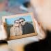 Visão em primeira pessoa de alguém segurando um porta-retrato de madeira com a foto de um casal sorridente à beira de um lago. O foco está na moldura, enquanto o fundo permanece desfocado.