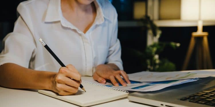 Close das mãos de uma revisora de textos escrevendo anotações em um caderno, com folhas de rascunho e notebook sobre a mesa em ambiente calmo.