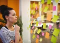 Mulher observando uma parede com post-its coloridos, refletindo sobre organização e planejamento no ambiente de trabalho.