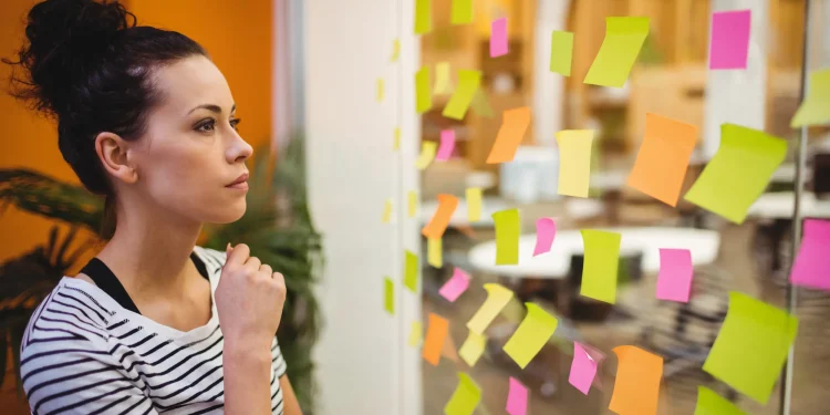 Mulher observando uma parede com post-its coloridos, refletindo sobre organização e planejamento no ambiente de trabalho.