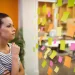 Mulher observando uma parede com post-its coloridos, refletindo sobre organização e planejamento no ambiente de trabalho.