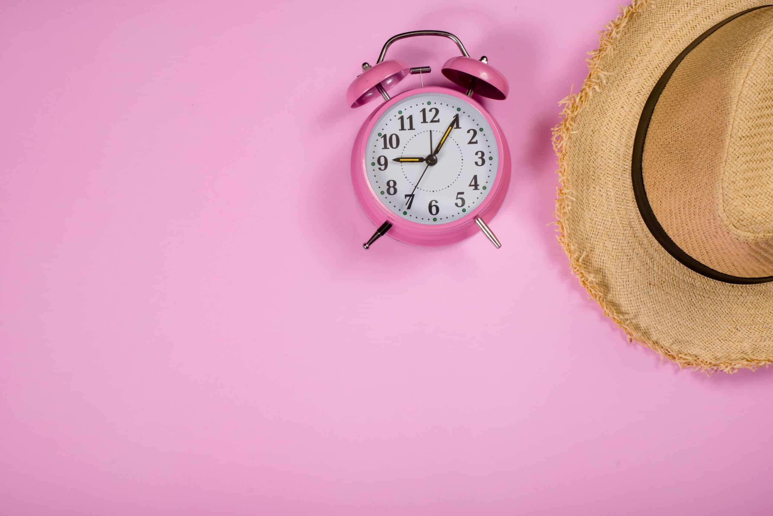 Visão superior de um despertador rosa analógico ao lado de um chapéu de palha sobre um fundo rosa sólido, representando o conceito de horário de verão e lazer.