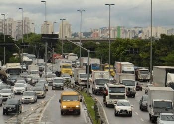 Vista aérea de uma rodovia movimentada com diversos carros, caminhões e vans em circulação sob um céu nublado, com prédios e vegetação ao fundo.