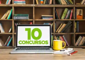 Laptop em mesa exibe “10 concursos” ao lado de caneca amarela e cadernos, com estante de livros ao fundo.