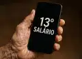 Mão segura celular que mostra a frase “13º salário” na tela em fundo escuro.