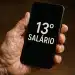 Mão segura celular que mostra a frase “13º salário” na tela em fundo escuro.