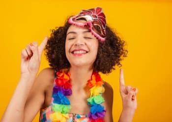 Uma mulher jovem sorridente, com cabelos cacheados, usa uma máscara de Carnaval rosa na testa e um colar de flores coloridas. Ela está com os olhos fechados e aponta os dedos indicadores para cima em um fundo amarelo vibrante.