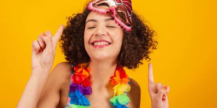 Uma mulher jovem sorridente, com cabelos cacheados, usa uma máscara de Carnaval rosa na testa e um colar de flores coloridas. Ela está com os olhos fechados e aponta os dedos indicadores para cima em um fundo amarelo vibrante.