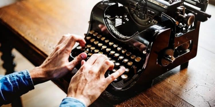 Close das mãos de uma pessoa utilizando uma máquina de escrever mecânica antiga de cor preta sobre uma mesa de madeira escura.